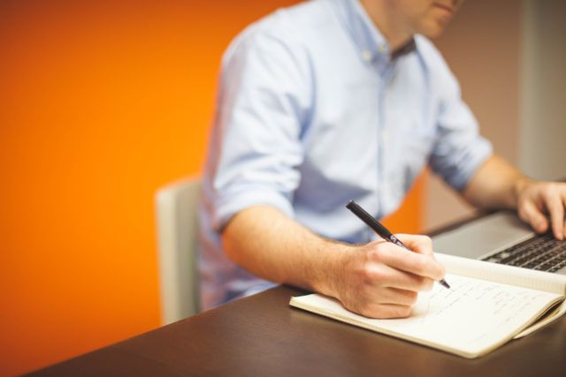 Concentrarsi in un mondo di notifiche: 6 abitudini pratiche A man sits in modern office, writing notes with a laptop open.