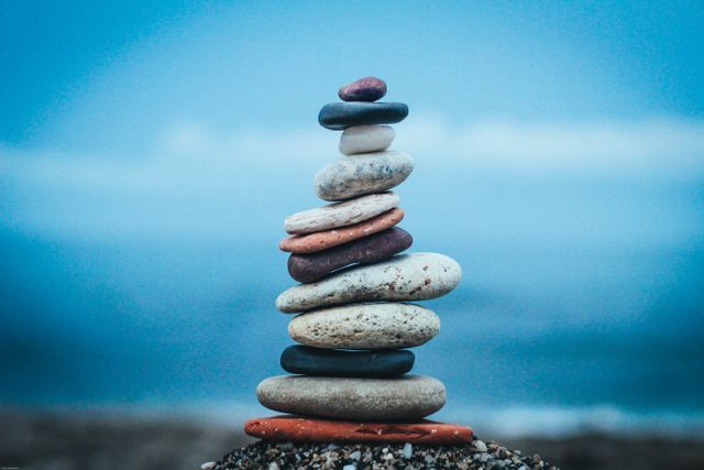Riscoprire il Tuo Equilibrio: Guida al Benessere Consapevole con ZENVIA.bio Closeup of stacked flat stones of various sizes and colors arranged on beach against blurred blue waving sea