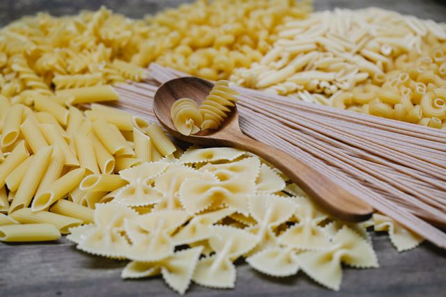 Pasta artigianale: storia, trafilatura al bronzo ed essiccazione lenta Arrangement of uncooked various pasta including spaghetti fusilli farfalle and penne heaped on table with wooden spoon